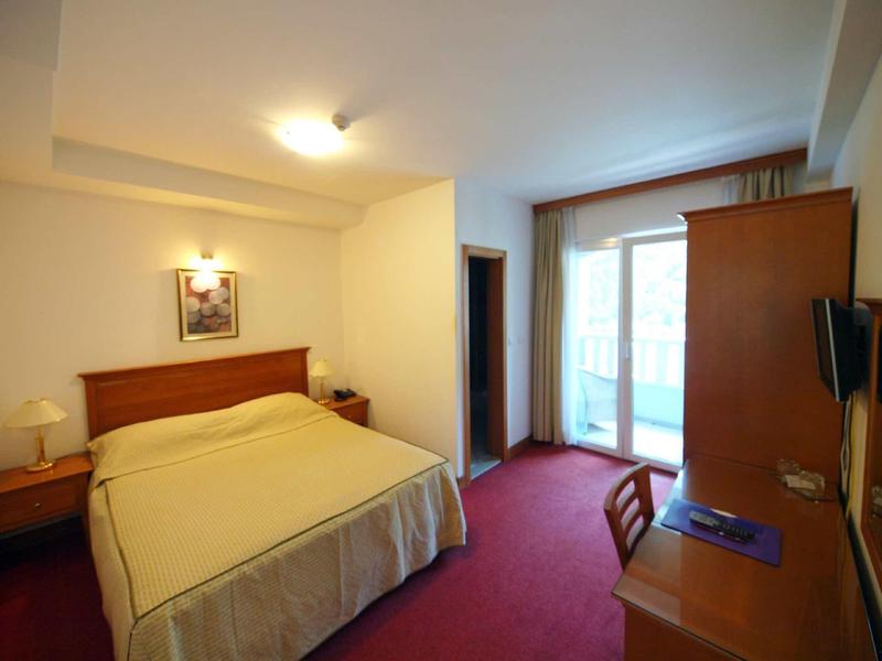 Einfaches Hotelzimmer mit Doppelbett, Schreibtisch, Stuhl, Tür zum Balkon und gedämpfter Beleuchtung.
