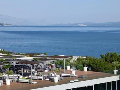 Dachterrasse mit Außenmöbeln und Meer im Hintergrund, Himmel klar, grüne Bäume am Rand.