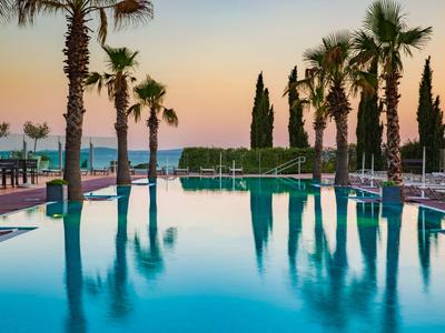 Ein glatter, blauer Hotelpool umgeben von Palmen und einer Terrasse mit Blick auf das Meer bei Sonnenuntergang.