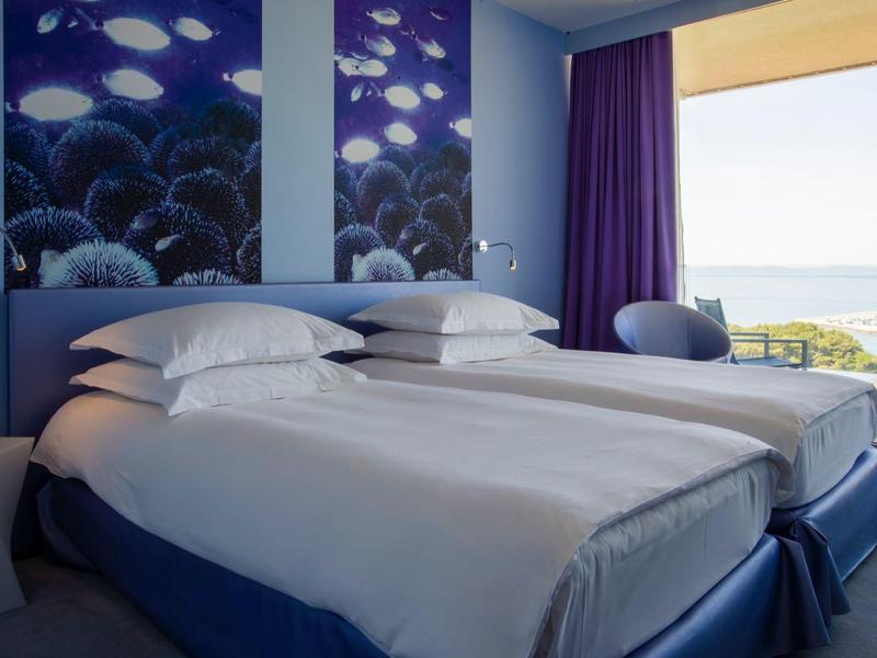 Camera d'hotel moderna con due letti, decorazione murale blu e balcone con vista mare.