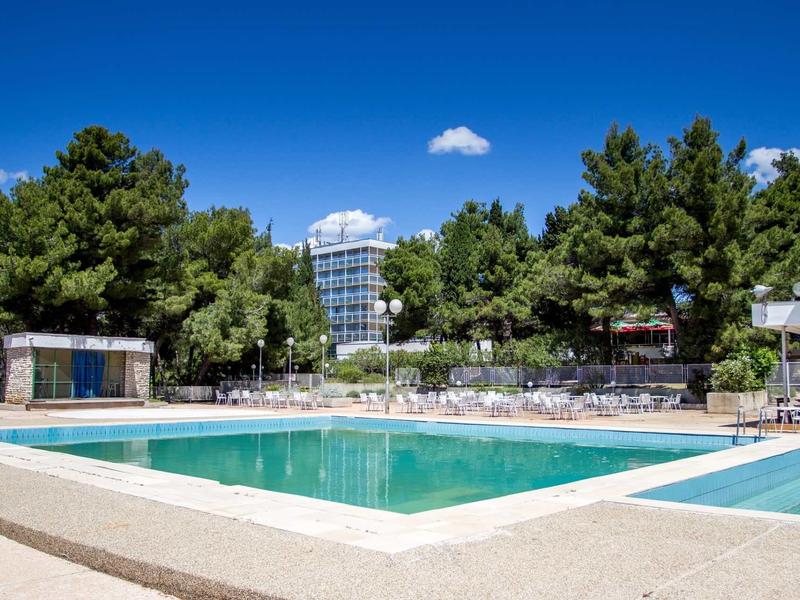 Piscina limpida con lettini circondata da pini sotto un cielo azzurro.