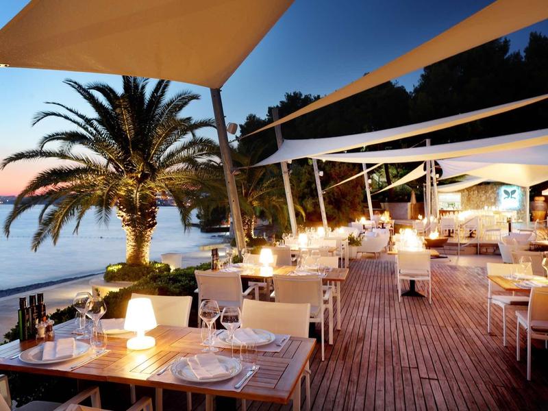 Večeře na terase u moře s palmami a teplým osvětlením pod plachtovými stříškami.