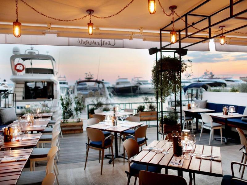 Moderní restaurace s dřevěnými stoly, židlemi a výhledem na přistavené lodě v přístavu.