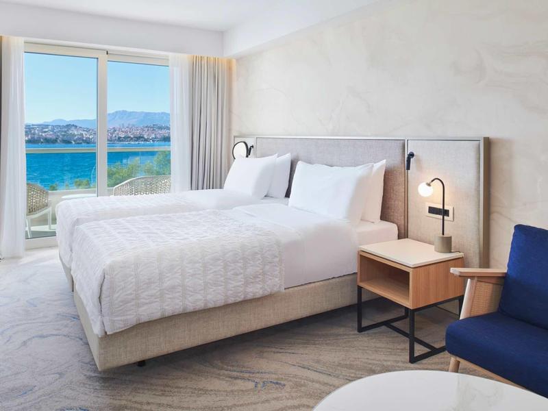 Moderní hotelový pokoj s manželskou postelí, výhledem na moře a útulnou sedací částí.