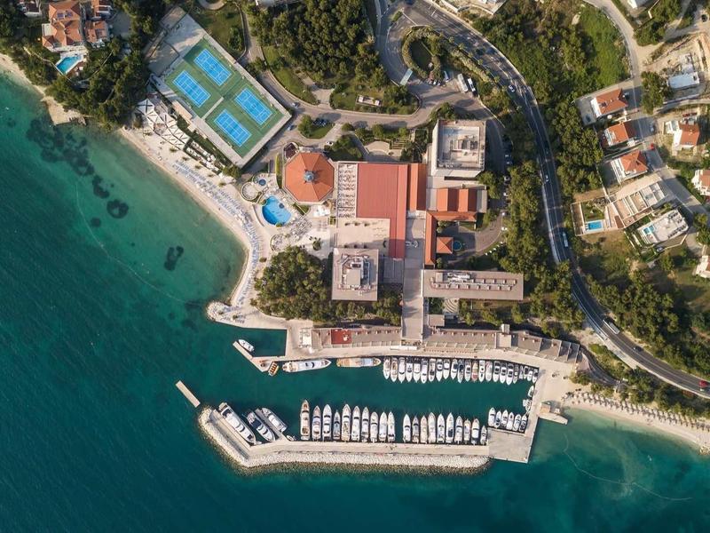 Letecký pohled na pobřežní hotel s bazénem, přístavem a několika loděmi ve čisté vodě.