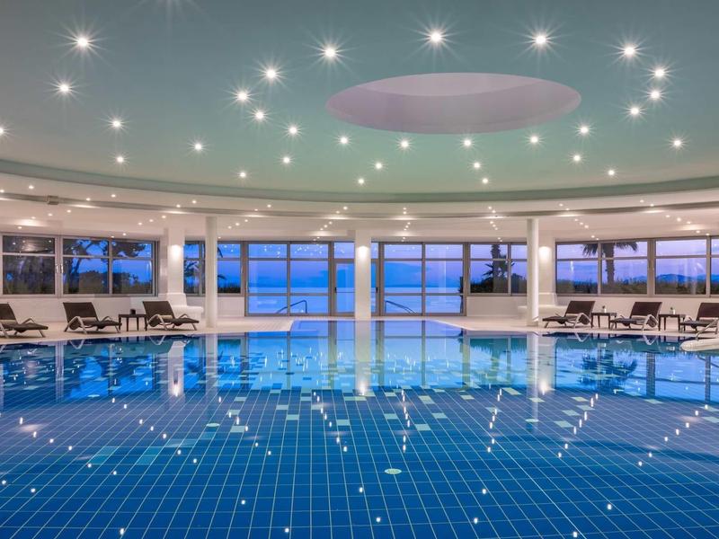 Velký vnitřní bazén s modrými dlaždicemi a lehátky při západu slunce.