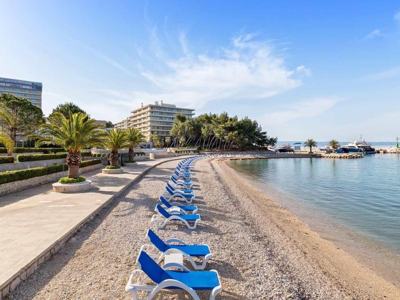 Prázdná pláž s modrými lehátky a hotelovými budovami v pozadí pod jasnou oblohou.