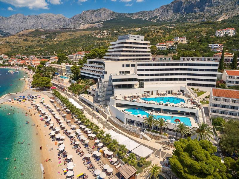 Großes Hotel mit Pool direkt am Strand und Bergblick unter einem blauen Himmel.