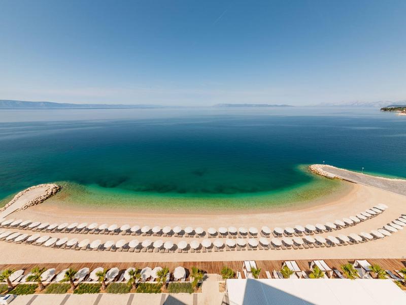 Strand mit Reihen von Liegestühlen und Schirmen vor ruhigem blauem Meer bei klarem Himmel.
