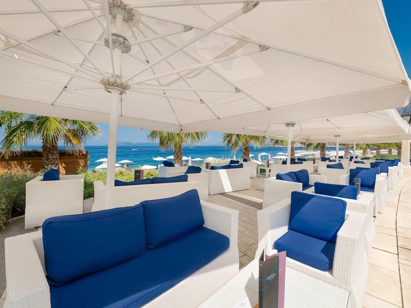 Außenlounge mit weißen Möbeln, blauen Kissen und Sonnenschirmen am Meer.