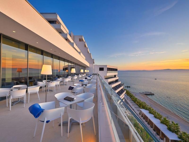 Moderne Hotelterrasse mit Meerblick und Sonnenuntergang, weißes Mobiliar und Glasgeländer.