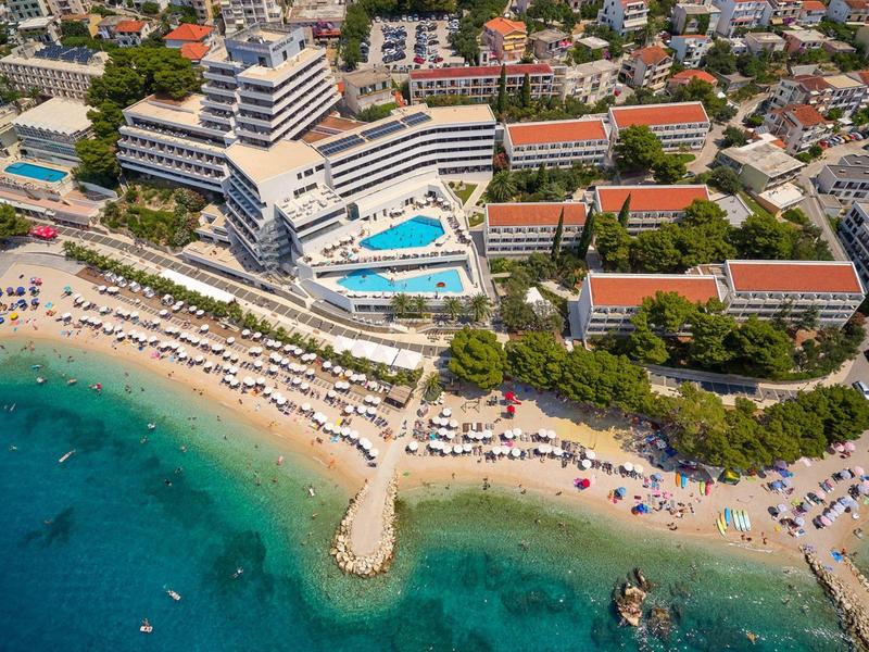 Hotel mit Pool und Liegestühlen am Sandstrand neben klarem Meerwasser in einer belebten Küstenstadt.