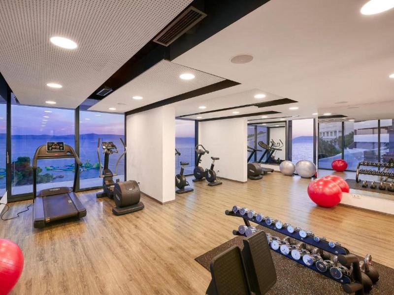 Helles Fitnessstudio mit Holzfußboden, Geräten und großen Fenstern mit Meerblick.