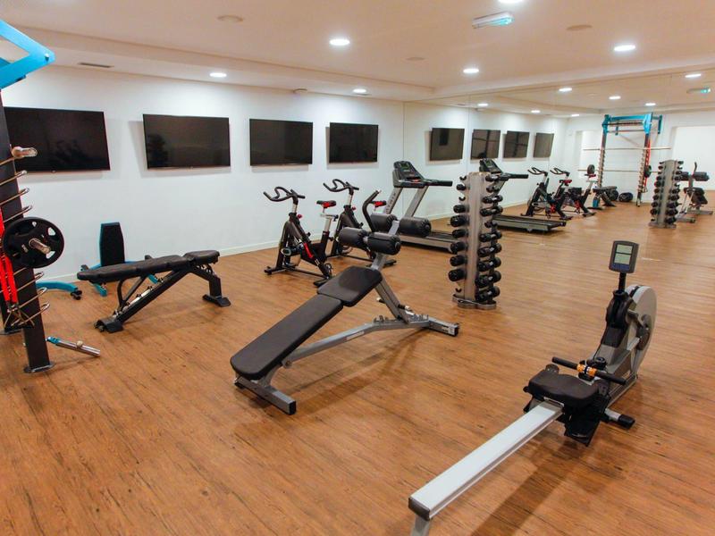 Salle de sport lumineuse avec sol en bois, divers équipements d'exercice et plusieurs téléviseurs au mur.