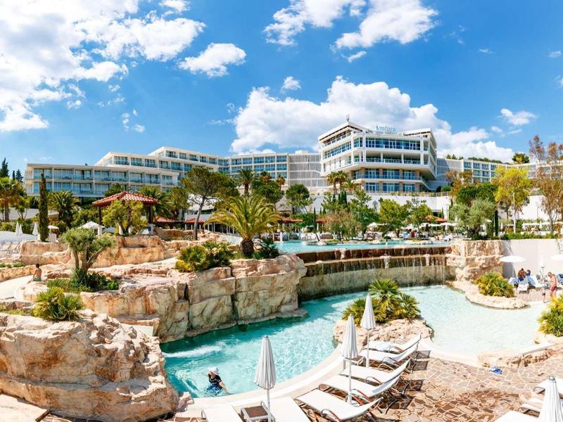 Station avec piscine, chaises longues, palmiers et bâtiments modernes sous un ciel ensoleillé.