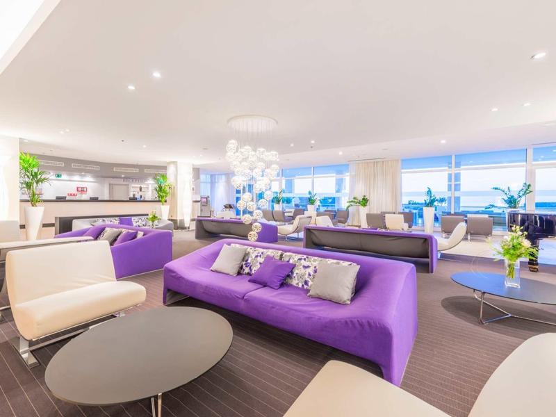 Espace lounge d'hôtel moderne avec canapés violet, fauteuils clairs et vue sur la mer.