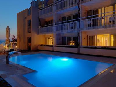 Piscina moderna de hotel al anochecer con agua iluminada y habitaciones con balcón al fondo.