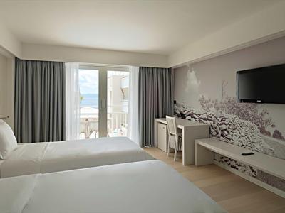 Habitación de hotel luminosa con dos camas, balcón y vista al mar a través de las ventanas.