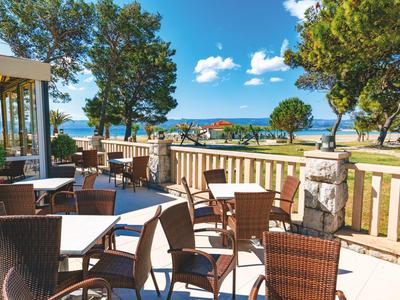 Terrasse mit braunen Stühlen und Tischen, Blick auf Bäume und das Meer bei klarem Himmel.
