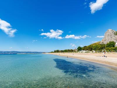 Klarer Strand mit türkisfarbenem Wasser, Sand und felsigem Hügel unter blauem Himmel.