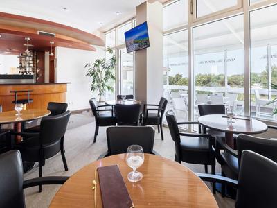 Moderner Hotel-Loungebereich mit Tischen, Stühlen und Bar in einem hellen Raum mit großen Fenstern.