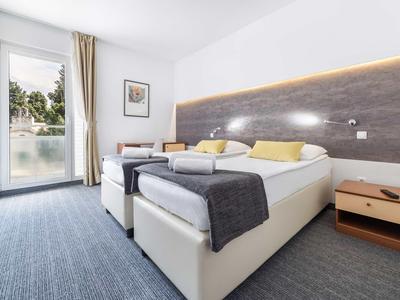 Nowoczesny pokój hotelowy z łóżkiem podwójnym, stolikiem nocnym i balkonem z widokiem na zieleń.