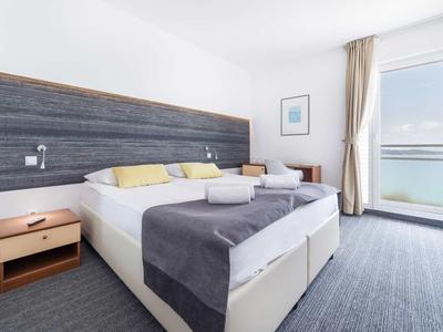 Jasny pokój hotelowy z podwójnym łóżkiem, szarą pościelą, szafką nocną i balkonem z widokiem na morze.