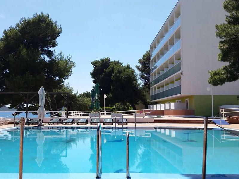 Ogrodzony basen na zewnątrz hotelu z leżakami i drzewami w tle pod niebieskim niebem.