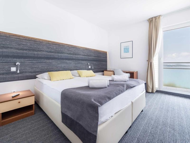 Jasny pokój hotelowy z podwójnym łóżkiem, szarą pościelą, szafką nocną i balkonem z widokiem na morze.