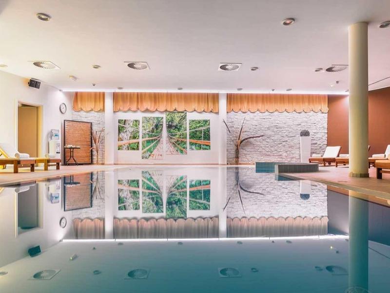 Jasny kryty basen z spokojną wodą, białymi filarami i przytulnymi miejscami do siedzenia.