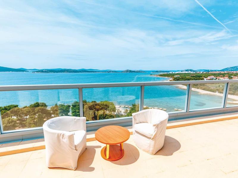 Balkon z dwoma białymi fotelami, okrągłym drewnianym stolikiem i widokiem na niebieskie morze i wybrzeże.