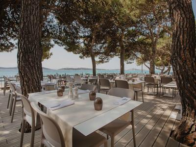 Freiluftrestaurant mit weißen Tischen unter schattigen Pinien am Meer