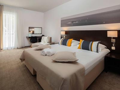 Modernes Hotelzimmer mit großem Doppelbett, bunten Kissen und Sitzbereich neben Fenster mit Vorhängen.