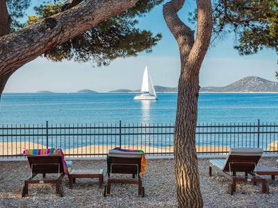 Blick durch Bäume auf drei Sonnenliegen am Kiesstrand mit Segelboot und Meer im Hintergrund.