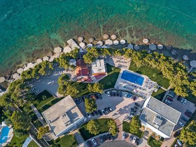 Luftaufnahme einer Küstenanlage mit Gebäuden, Garten, Pool und türkisblauem Wasser.