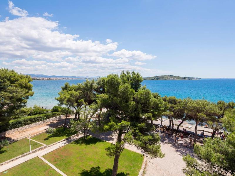 Grüne Bäume und Wege an einer Küste mit blauem Meer und bewölktem Himmel im Hintergrund.