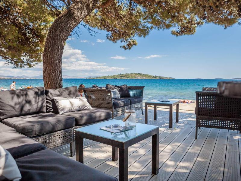 Gemütliche Sitzgruppe mit braunen Polstern auf Holzdeck unter Pinienbaum am blauen Meer.