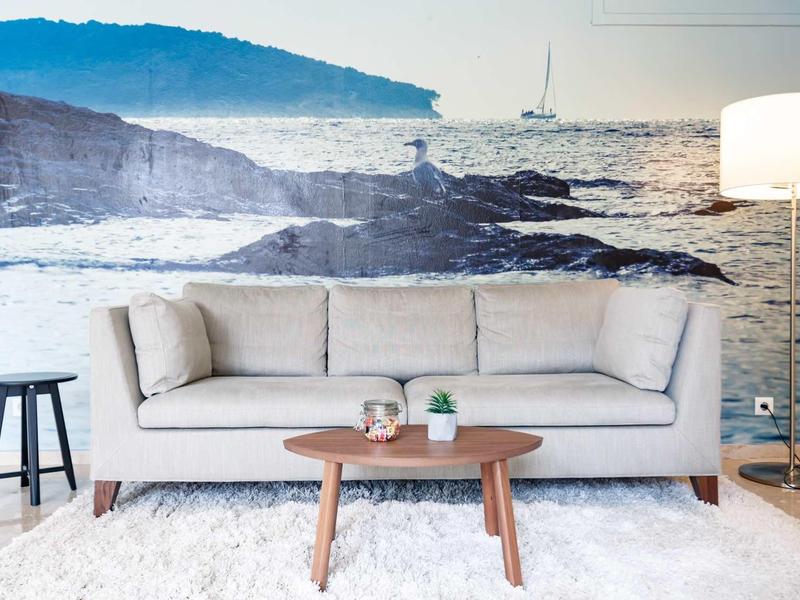 Wohnzimmer mit hellgrauem Sofa, kleinem Holztisch, weißem Teppich und Wandbild einer Küstenlandschaft.