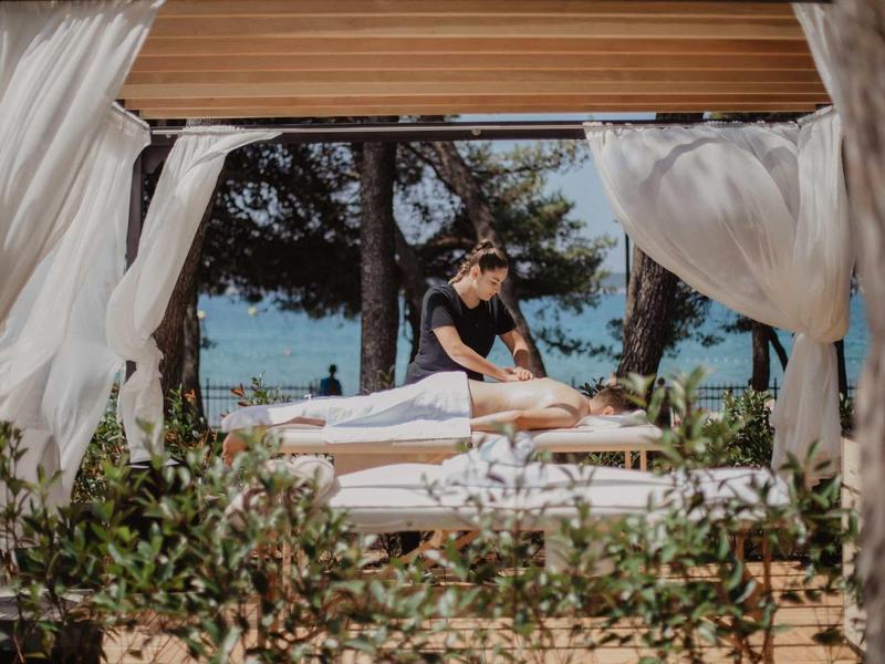 Frau erhält Massage auf Liege im Freien, umgeben von weißen Vorhängen und Bäumen am Meer.