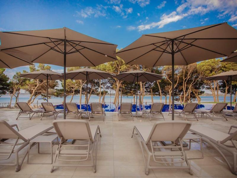 Liegestühle mit blauen Polstern und großen Sonnenschirmen vor Bäumen und blauem Himmel.