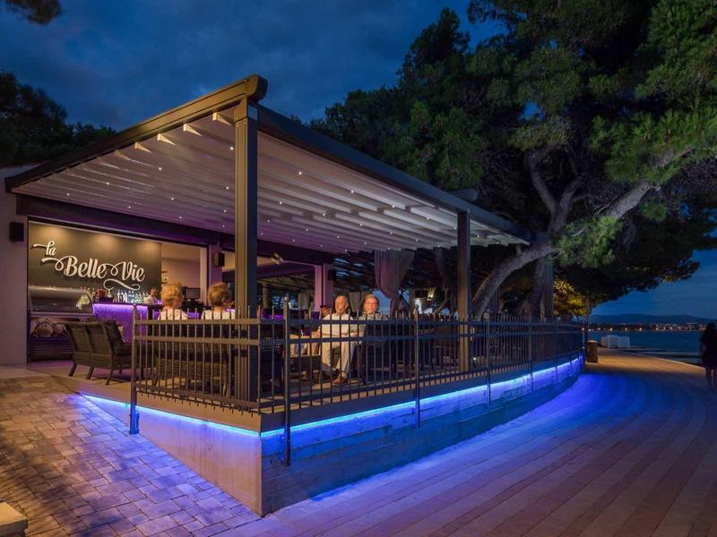 Beleuchtetes Restaurant mit überdachtem Sitzbereich und Holzterrasse am Abend, umgeben von Bäumen.