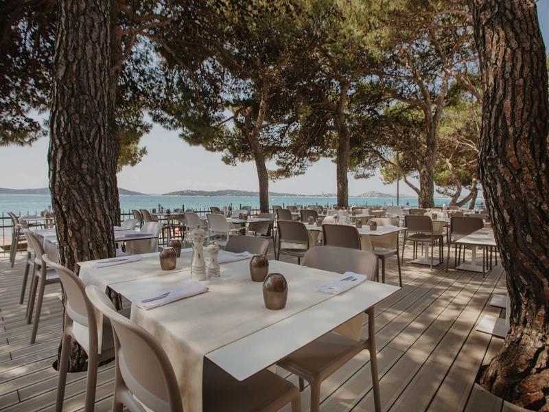 Freiluftrestaurant unter Bäumen mit Tischen, Stühlen und Blick auf das Meer an einem sonnigen Tag.