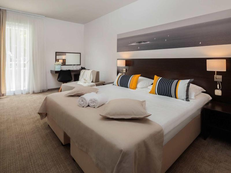 Modernes Hotelzimmer mit großem Doppelbett, bunten Kissen und Sitzbereich neben Fenster mit Vorhängen.