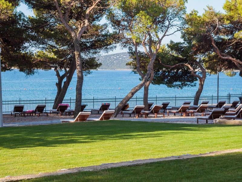 Grüne Rasenfläche mit Liegestühlen und Bäumen, dahinter Blick aufs blaue Meer.