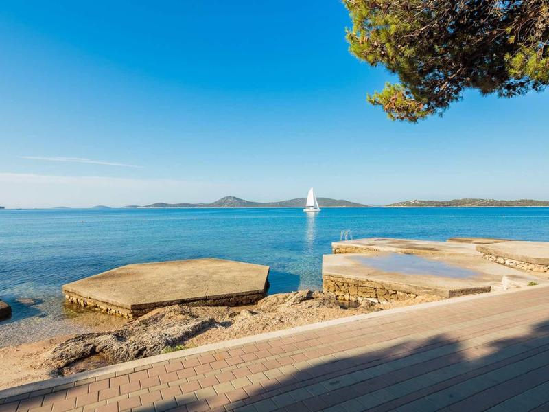 Klarer blauer Himmel über ruhigem Meer mit Segelboot und steinigen Küstenflächen im Vordergrund.