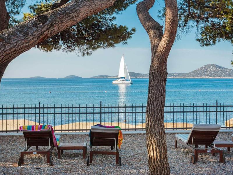 Blick durch Bäume auf drei Sonnenliegen am Kiesstrand mit Segelboot und Meer im Hintergrund.