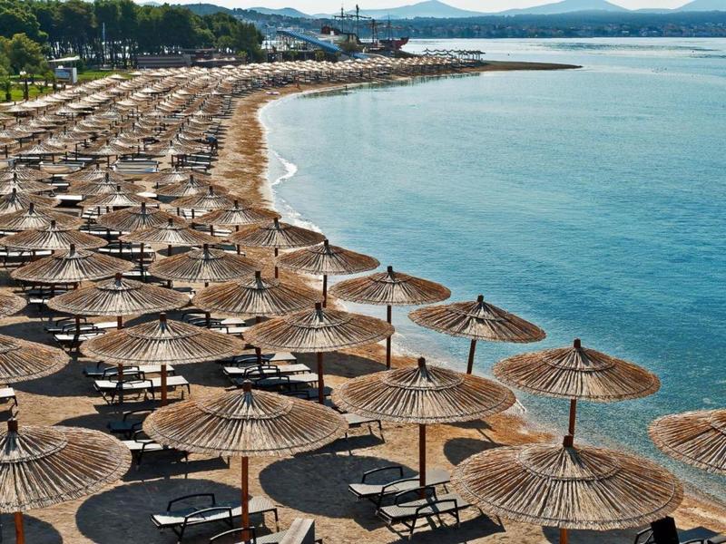Lange zandstrand met veel strooien parasols en ligstoelen naast kalm blauw water.