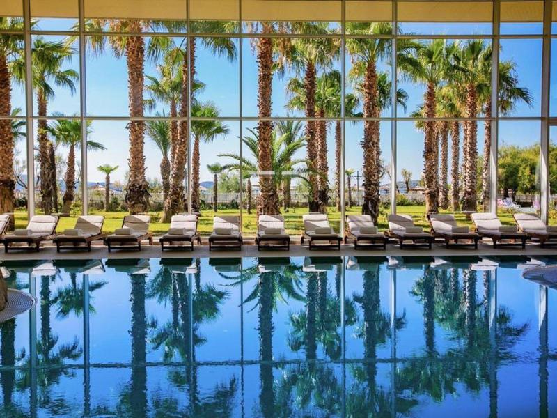 Binnenzwembad met ligstoelen en palmbomen achter grote ramen op een zonnige dag.
