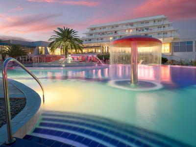 Atmosfera serale alla piscina dell'hotel illuminata con giochi d'acqua e palme.