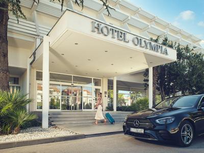Entrance area of Hotel Olympia with a black car and plants on a sunny day.